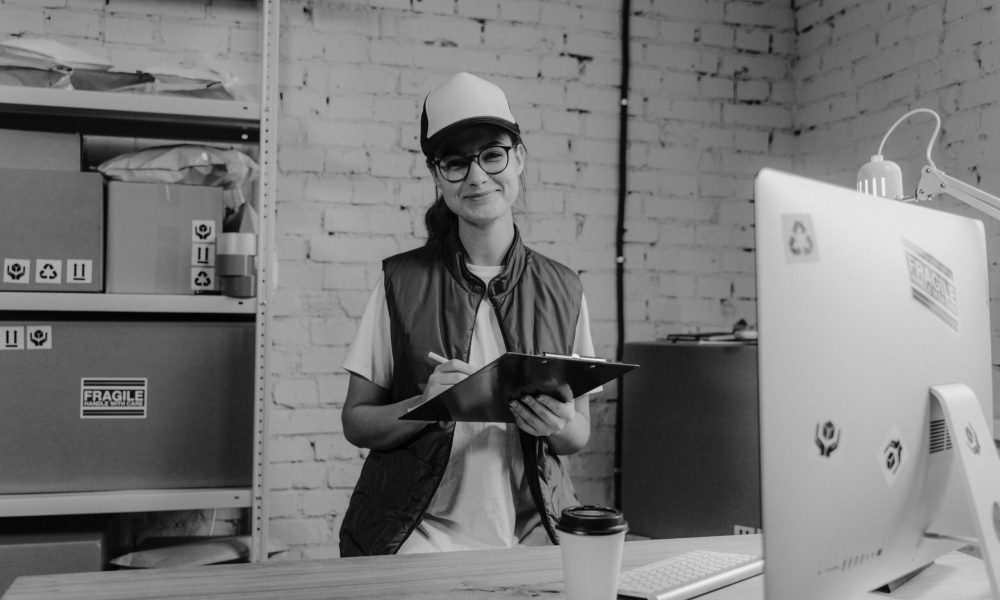 A smiling woman in a logistics office holding a clipboard, wearing casual attire.